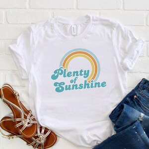 May include: White t-shirt with the text "Plenty of Sunshine" in a retro font, featuring a rainbow design. The image also includes blue jeans and brown sandals with gold accents.