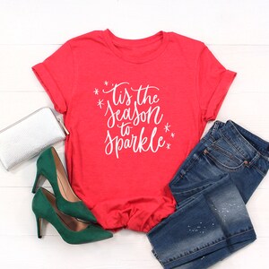 May include: Red t-shirt with white text that says "Tis the season to sparkle". The shirt is laying on a white wooden surface with a pair of green high heels, a silver clutch, and blue jeans.