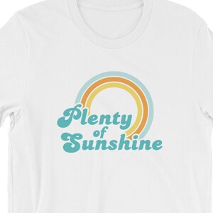 May include: White t-shirt with a retro design. The shirt features the text "Plenty of Sunshine" in a teal, stylized font, with a rainbow graphic in orange, yellow, and teal. The t-shirt is made of a soft material.