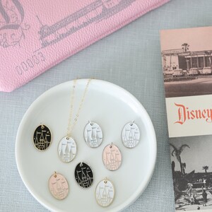 May include: A white ceramic plate with a gold chain necklace and seven oval-shaped charms. The charms are in different colors, including black, white, pink, and gold. Each charm features a white silhouette of Cinderella's castle with a gold outline.