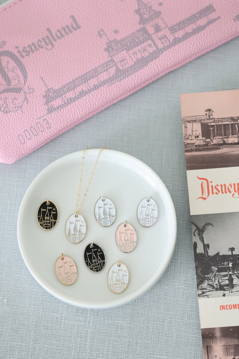 May include: A white ceramic plate with a gold chain necklace and seven oval-shaped charms. The charms are in different colors, including black, white, pink, and gold. Each charm features a white silhouette of Cinderella's castle with a gold outline.
