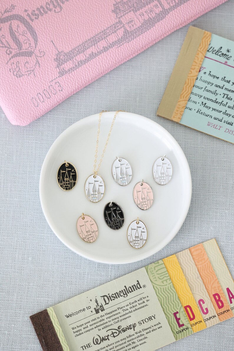 May include: A gold chain necklace with six different colored oval charms featuring a black and white outline of Cinderella's castle. The charms are displayed on a white plate. The text "Welcome to Disneyland" is visible on a ticket.