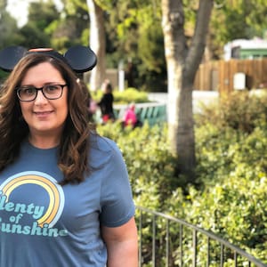 May include: A woman wearing a blue t-shirt with the text "Plenty of Sunshine" and a rainbow design. She is also wearing black glasses and Mickey Mouse ears. The background includes greenery and a blurred outdoor setting.