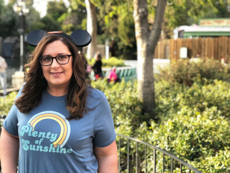 May include: A woman wearing a blue t-shirt with the text "Plenty of Sunshine" and a rainbow design. She is also wearing black glasses and Mickey Mouse ears. The background includes greenery and a blurred outdoor setting.