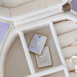 May include: Two rectangular pendants with a gold border and a Mickey Mouse design are displayed inside a cream-colored jewelry box. One pendant is light purple, the other is beige. The jewelry box has a gold zipper.
