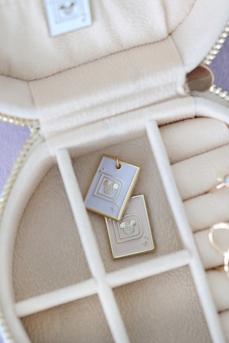 May include: Two rectangular pendants with a gold border and a Mickey Mouse design are displayed inside a cream-colored jewelry box. One pendant is light purple, the other is beige. The jewelry box has a gold zipper.