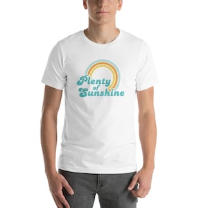 May include: White t-shirt with the text "Plenty of Sunshine" in a retro font, above a rainbow design in shades of orange, teal, and yellow. The shirt is short-sleeved and worn by a person.