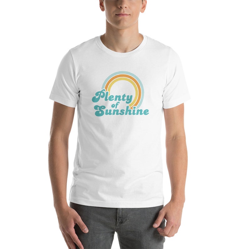 May include: White t-shirt with the text "Plenty of Sunshine" in a retro font, above a rainbow design in shades of orange, teal, and yellow. The shirt is short-sleeved and worn by a person.