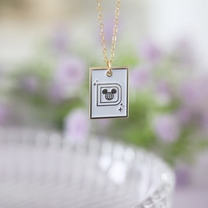May include: Gold-colored chain necklace with a white rectangular pendant. The pendant features a black and white graphic of a stylized letter "D" with a globe and Mickey Mouse ears. The necklace is displayed against a blurred floral background.
