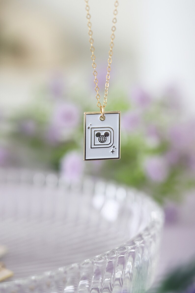 May include: Gold-colored chain necklace with a white rectangular pendant. The pendant features a black and white graphic of a stylized letter "D" with a globe and Mickey Mouse ears. The necklace is displayed against a blurred floral background.