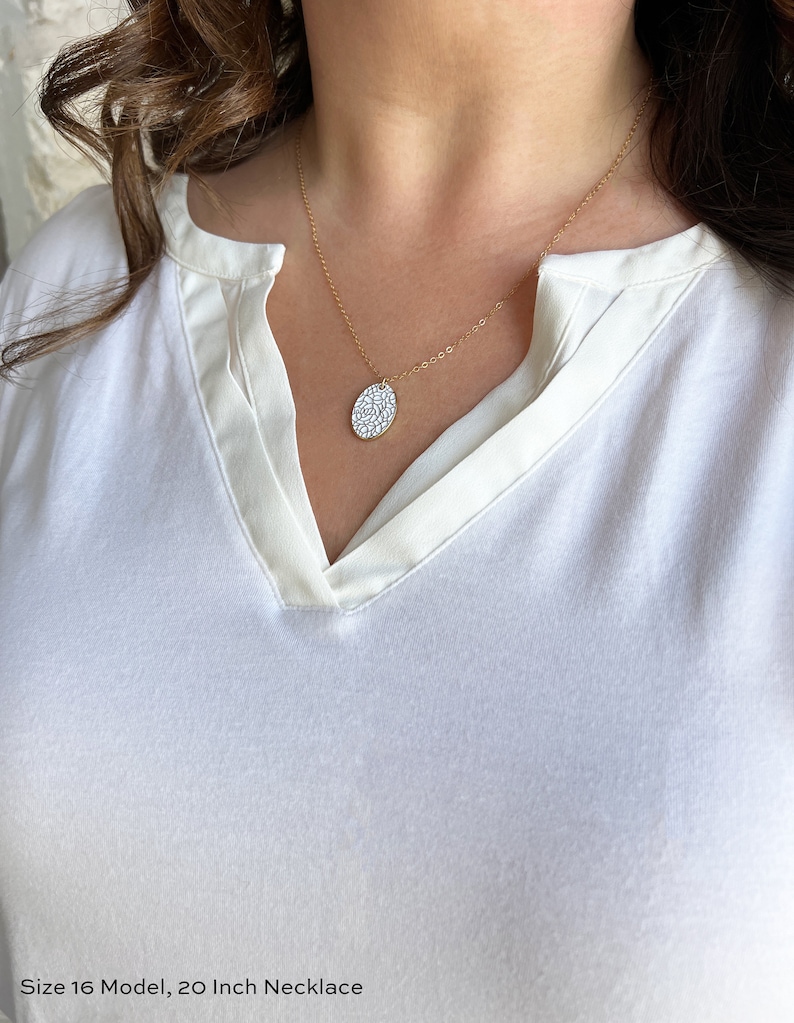 May include: A gold chain necklace with a white oval pendant featuring a delicate floral pattern. The necklace is 20 inches long.