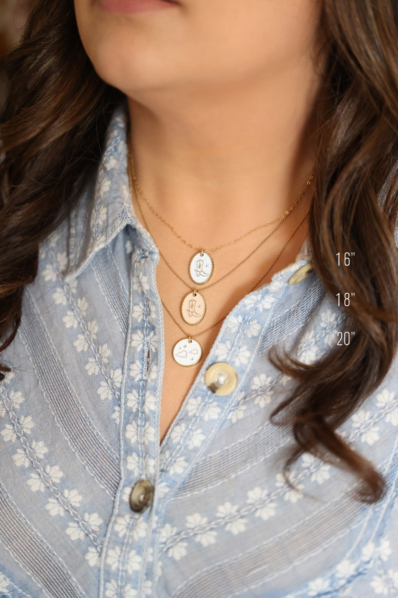 May include: Three gold necklaces of varying lengths, 16, 18, and 20 inches, each with a different pendant. The pendants feature engraved designs of a cowboy boot, a cowboy boot, and a cloud with stars. The necklaces are displayed against a light blue floral shirt.