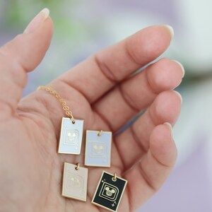 May include: A gold chain necklace with four rectangular pendants in white, lavender, pink, and black. Each pendant features a gold-colored design of a cartoon character's face. The pendants are held in a person's open hand.