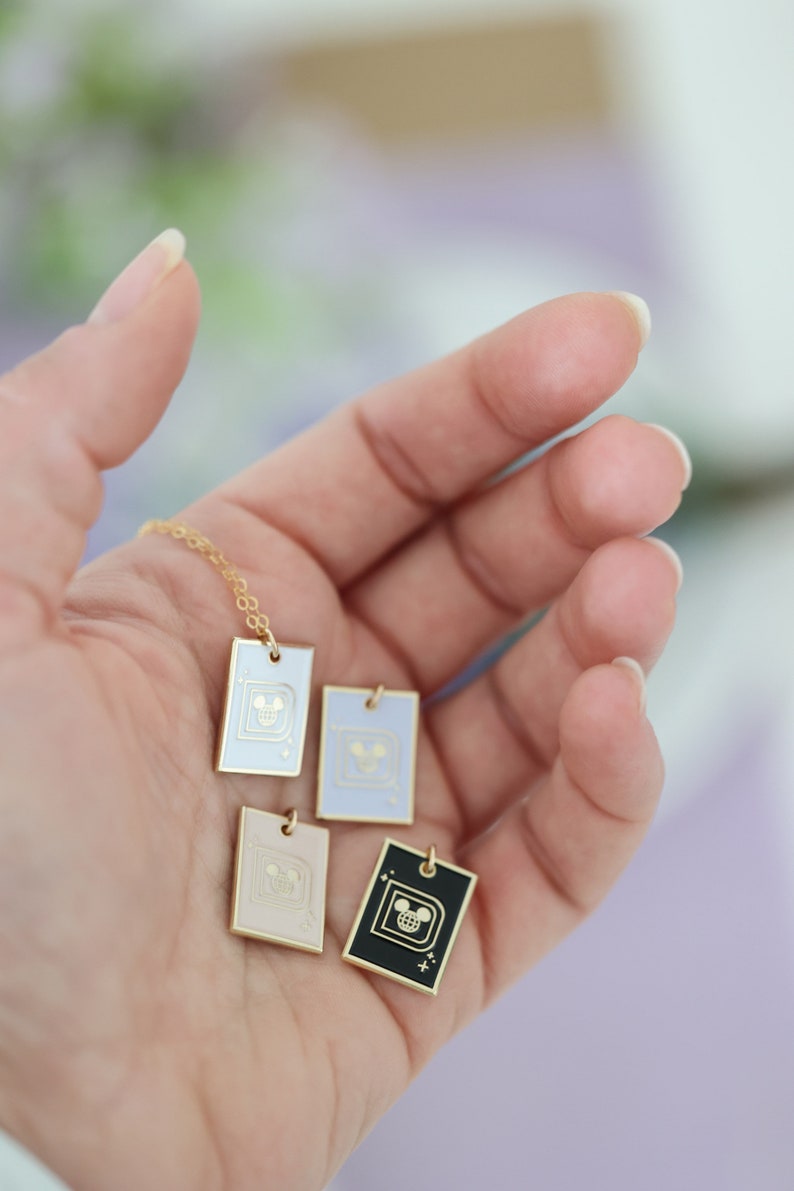 May include: A gold chain necklace with four rectangular pendants in white, lavender, pink, and black. Each pendant features a gold-colored design of a cartoon character's face. The pendants are held in a person's open hand.