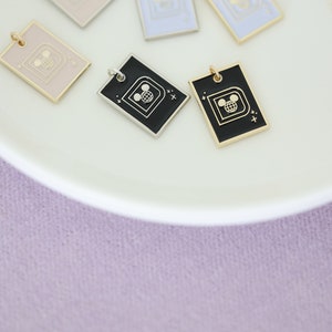 May include: Rectangular enamel pendants in various pastel colors, including pink, blue, and black, with gold or silver-tone borders. Each pendant features a Mickey Mouse head and globe design. The pendants are displayed on a white plate against a soft purple background.