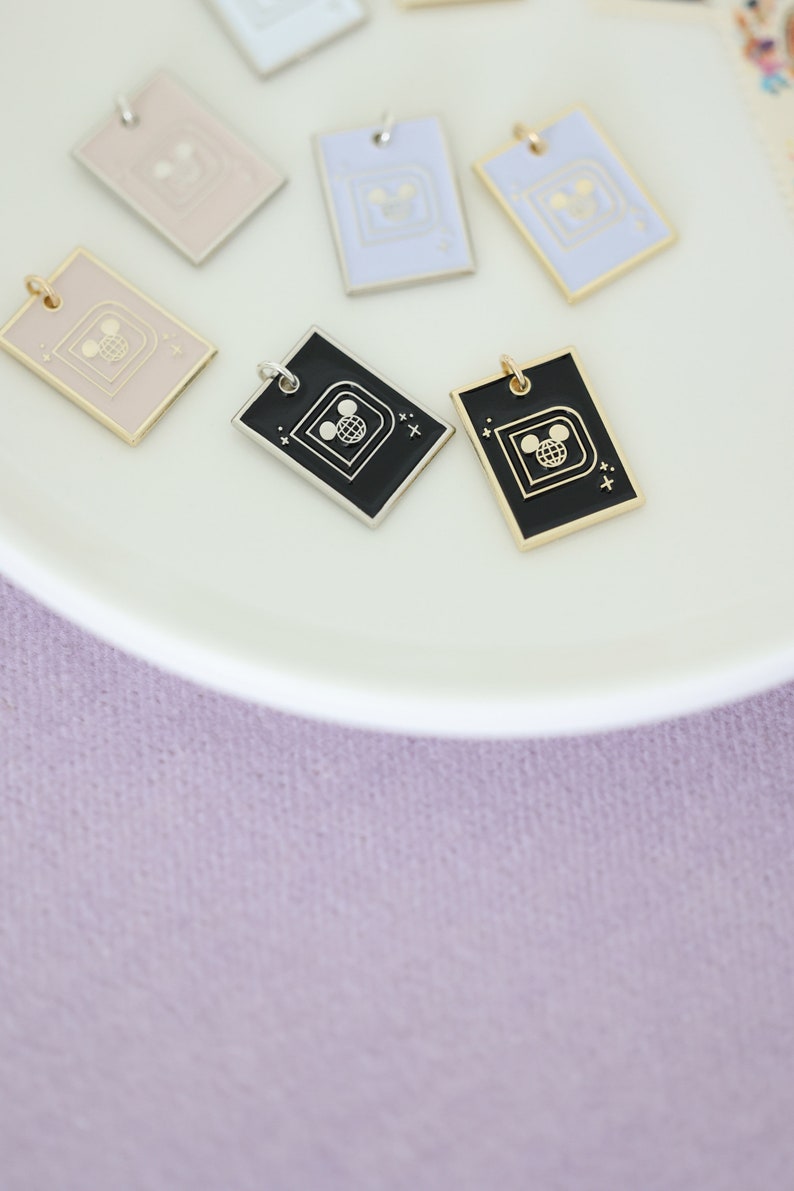 May include: Rectangular enamel pendants in various pastel colors, including pink, blue, and black, with gold or silver-tone borders. Each pendant features a Mickey Mouse head and globe design. The pendants are displayed on a white plate against a soft purple background.