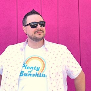 May include: A person wearing a white t-shirt with the text "Plenty of Sunshine" and a button-down shirt with a flamingo pattern. The person is wearing black sunglasses and a black watch. The background is a bright pink wall.