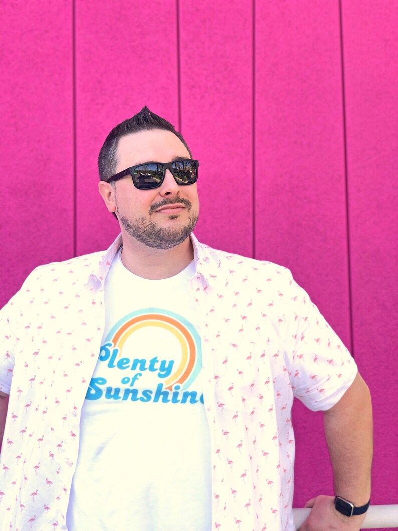 May include: A person wearing a white t-shirt with the text "Plenty of Sunshine" and a button-down shirt with a flamingo pattern. The person is wearing black sunglasses and a black watch. The background is a bright pink wall.