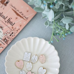 May include: A white ceramic plate with a scalloped edge holds eight charms featuring a white castle with pink accents. The charms are in various shapes, including hearts, ovals, and squares.