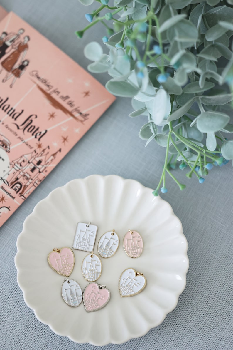 May include: A white ceramic plate with a scalloped edge holds eight charms featuring a white castle with pink accents. The charms are in various shapes, including hearts, ovals, and squares.