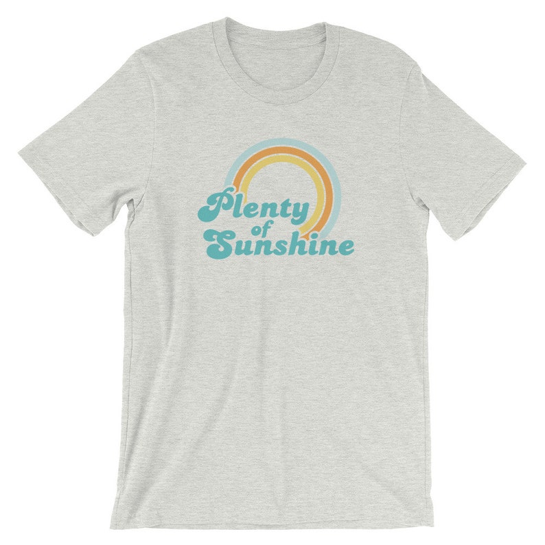 May include: A light gray t-shirt with a crew neck. The shirt features the text "Plenty of Sunshine" in a retro font, with a rainbow design in shades of orange, yellow, and teal.