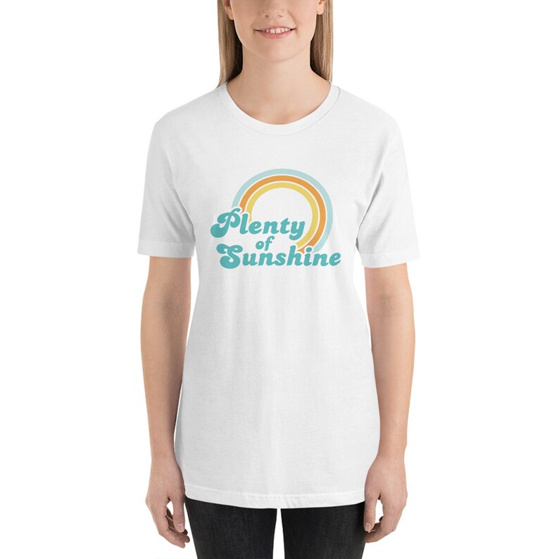 May include: White t-shirt with the text "Plenty of Sunshine" in a retro font, above a rainbow design in shades of blue, orange, and yellow. The shirt is short-sleeved and appears to be made of a soft material.