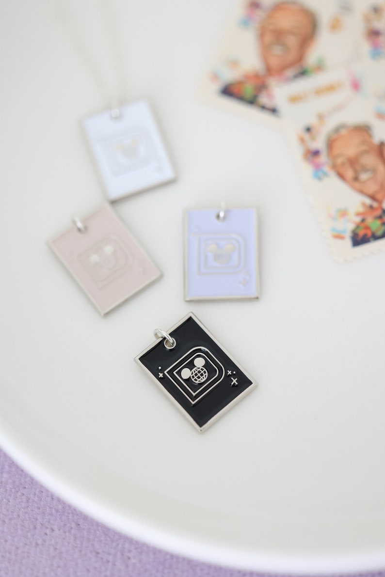 May include: Four rectangular pendant necklaces in white, pink, lavender, and black, each with a silver border and a Disney-themed design. The black pendant features a Mickey Mouse globe design. Stamps with a man's portrait are in the background.