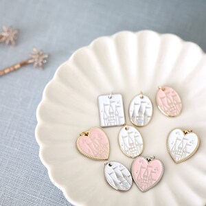 May include: A collection of seven pink and white enamel charms featuring a castle design. The charms are in the shapes of a square, oval, and heart. The charms are displayed on a white plate with a scalloped edge.