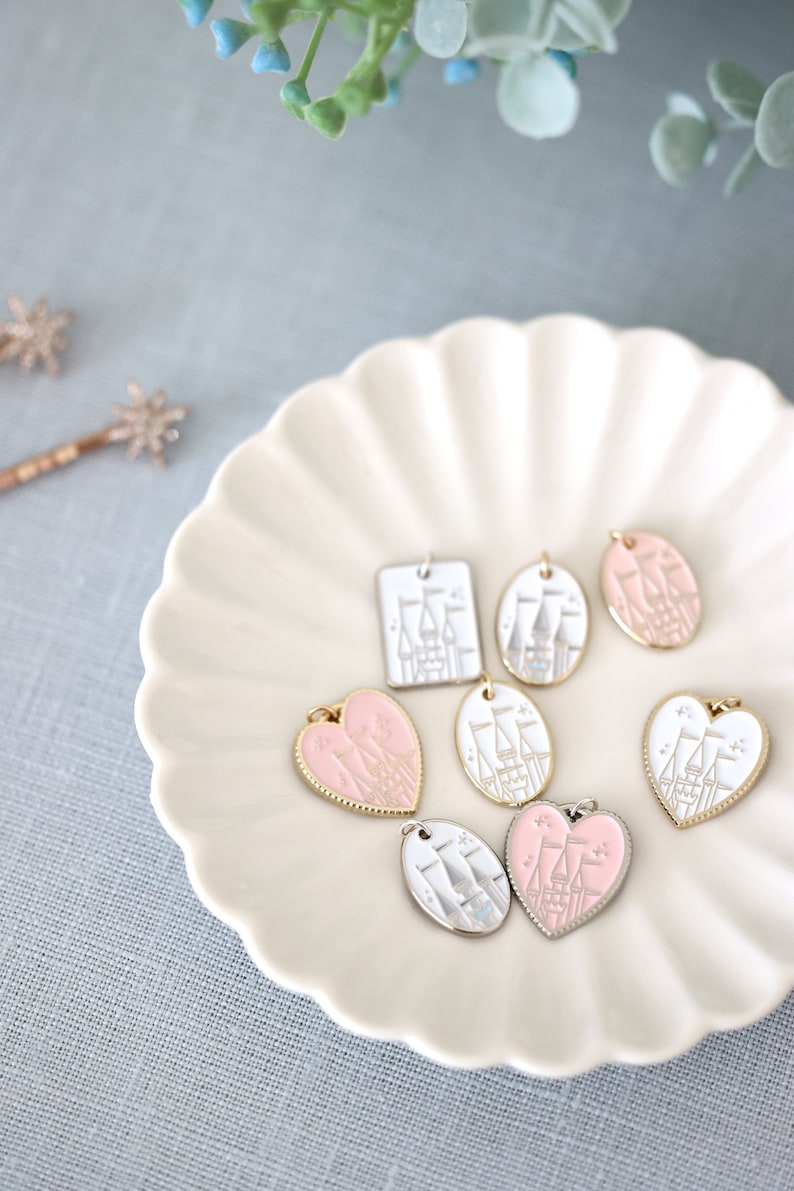 May include: A collection of seven pink and white enamel charms featuring a castle design. The charms are in the shapes of a square, oval, and heart. The charms are displayed on a white plate with a scalloped edge.