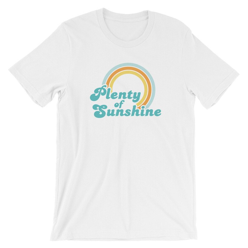 May include: White t-shirt with a retro design. The shirt features the text "Plenty of Sunshine" in a teal, stylized font, with a rainbow graphic in orange, yellow, and teal. The t-shirt is made of a soft material.
