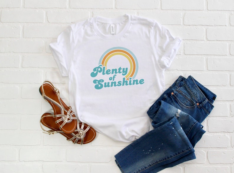 May include: White t-shirt with the text "Plenty of Sunshine" in a retro font, featuring a rainbow design. The image also includes blue jeans and brown sandals with gold accents.