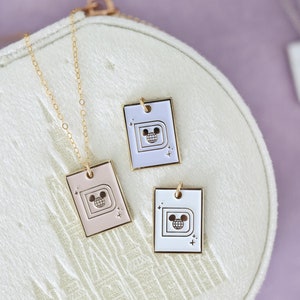 May include: Three rectangular pendant necklaces with a gold chain. The pendants are in pink, white, and lavender, each featuring a gold outline of a "D" with a Mickey Mouse head inside a globe. The pendants are on a white, textured surface.