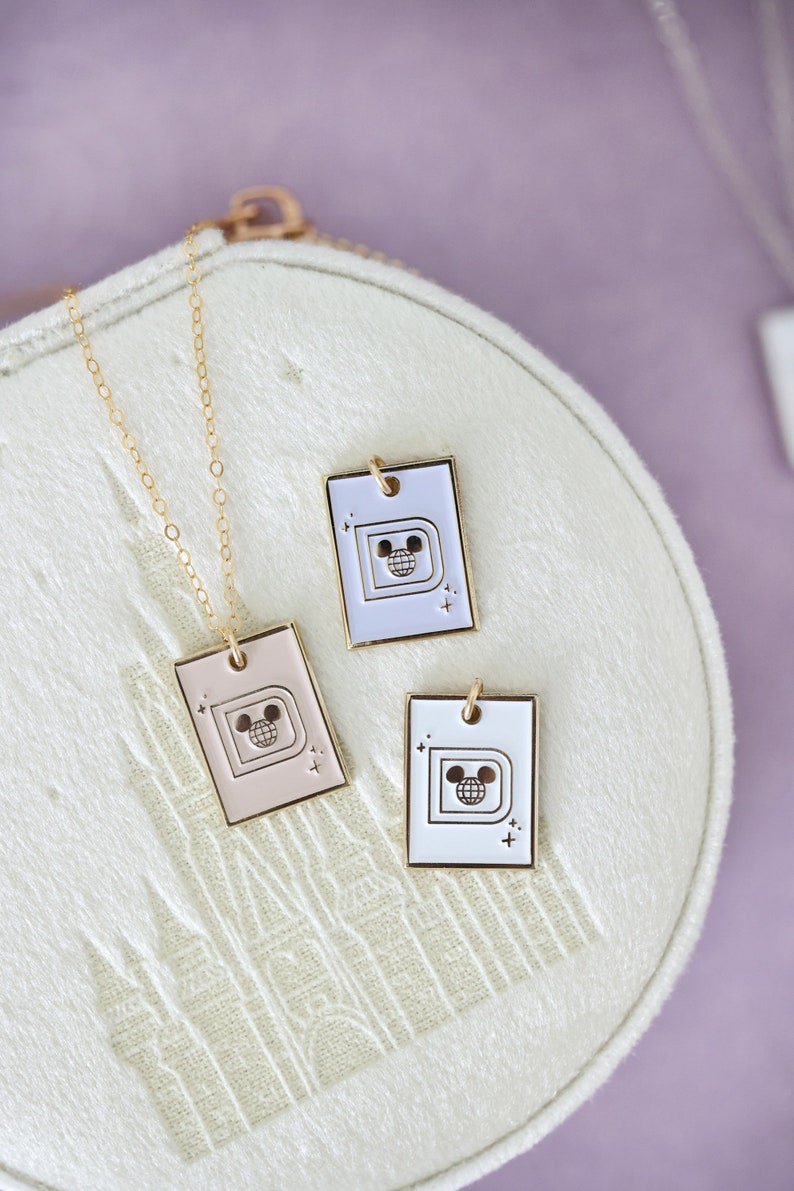 May include: Three rectangular pendant necklaces with a gold chain. The pendants are in pink, white, and lavender, each featuring a gold outline of a "D" with a Mickey Mouse head inside a globe. The pendants are on a white, textured surface.