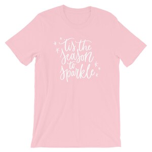 May include: Pink t-shirt with white text that reads "Tis the season to sparkle" with snowflakes on either side of the text.