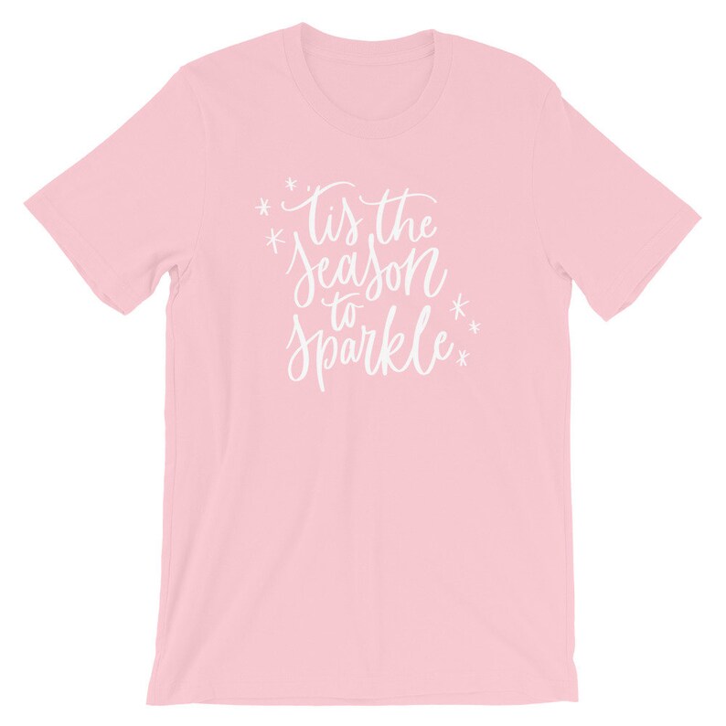 May include: Pink t-shirt with white text that reads "Tis the season to sparkle" with snowflakes on either side of the text.