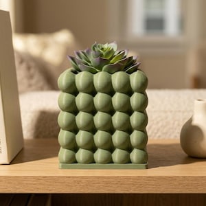 May include: A sage green square planter with a textured bubble surface pattern. The planter features a grid of rounded protrusions and contains a faux succulent plant with green and purple-tipped leaves, resting on a flat square base.