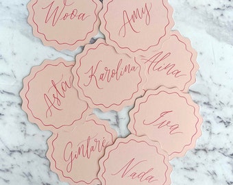 May include: Pink scalloped place cards with names written in pink calligraphy. The names are: Wooa, Amy, Alina, Karolina, Asta, Ieva, Gintare, and Nada.