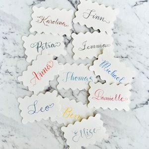 May include: Ten place cards with different names written in calligraphy. The names are: Karolina, Petra, Finn, Jemma, Anna, Thomas, Michael, Danielle, Leo, Ben, and Elise.