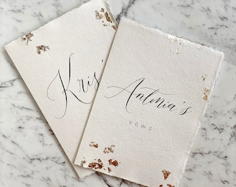 Puede incluir: Dos tarjetas blancas con motas doradas. La tarjeta superior tiene el texto "Kris'" escrito en cursiva. La tarjeta inferior tiene el texto "Antonia's Vows" escrito en cursiva.