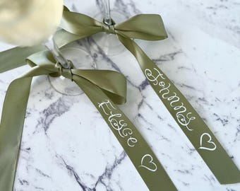 May include: Two olive green satin ribbons tied around champagne flutes with the names "Elyse" and "John" written in white calligraphy on the ribbons.