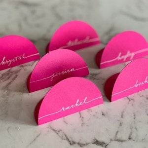 May include: A collection of six semi-circular hot pink place cards, each featuring a white horizontal line and a name written in white cursive script. The names visible on the cards include Krystal, Jessica, Rachel, Hayley, and Beck.