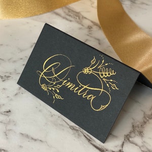 May include: A folded black place card featuring the name Dimitra written in elegant gold calligraphy. The script is accompanied by delicate gold botanical illustrations, creating a high-contrast design on the matte black cardstock surface.