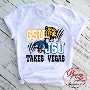 May include: White t-shirt with a graphic design. The design features the text "GSU vs JSU TAKES VEGAS", a yellow and black tiger head, a blue tiger head, and claw marks.