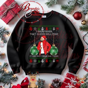 以下が含まれることがあります： 緑と赤のデザインの黒いクリスマスセーター。赤いジャケットとサンタの帽子をかぶった男性が描かれています。セーターのテキストは「I know when that sleigh bell ring it can only mean one thing」です。