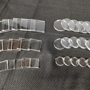 May include: A collection of clear acrylic game tokens, including square and circular shapes. The tokens are arranged on a dark, textured surface, suitable for tabletop games or crafting projects.