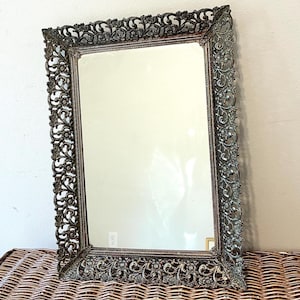 May include: An ornate, rectangular mirror with a decorative, dark metal frame. The frame features intricate floral and scrollwork designs. The mirror is displayed on a woven wicker surface, reflecting the neutral-colored wall behind it.