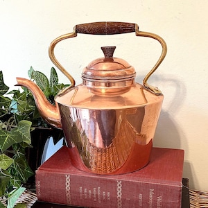 May include: A copper tea kettle with a wooden handle and brass accents. The kettle sits atop a stack of vintage books with red and brown covers. A green plant is visible on the left side of the image.
