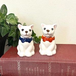 May include: Two white ceramic dog figurines sitting on top of two stacked books. One dog wears a dark blue bow tie, and the other wears a bright orange bow tie. Both dogs have painted brown noses, black eyes, and upright ears in a seated position.