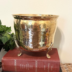 May include: A hammered brass planter with three decorative feet. The planter sits atop two antique books with red and black covers. The books are stacked on a woven wicker surface. Green foliage is visible on the left side.