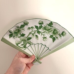 Op de afbeelding: Een decoratieve waaier in wit en groen, met een bloemenontwerp. De waaier heeft een groen bloemenpatroon op een witte achtergrond, met een groene rand. De waaier wordt tegen een witte muur gehouden.
